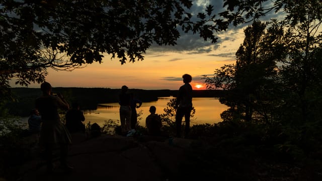 A group silhouetted on a rock outcrop above a lake watching the sun set, one person taking a photograph