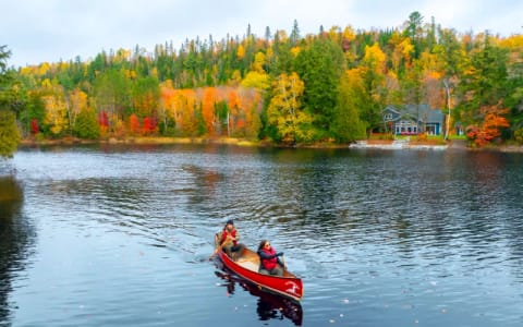 A red canoe crossing a lake below a shoreline of trees in full fall colour