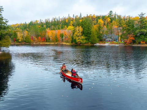 A red canoe crossing a lake below a shoreline of trees in full fall colour